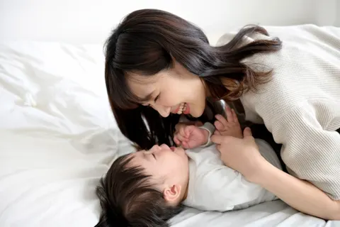 ベッドの上で、仰向けの赤ちゃんに顔を寄せてほほえむ女性