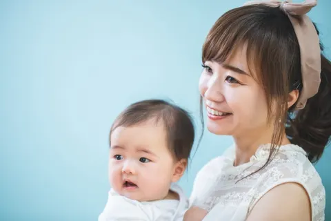 水色の無地の背景の前で、赤ちゃんを抱いてほほえむ女性